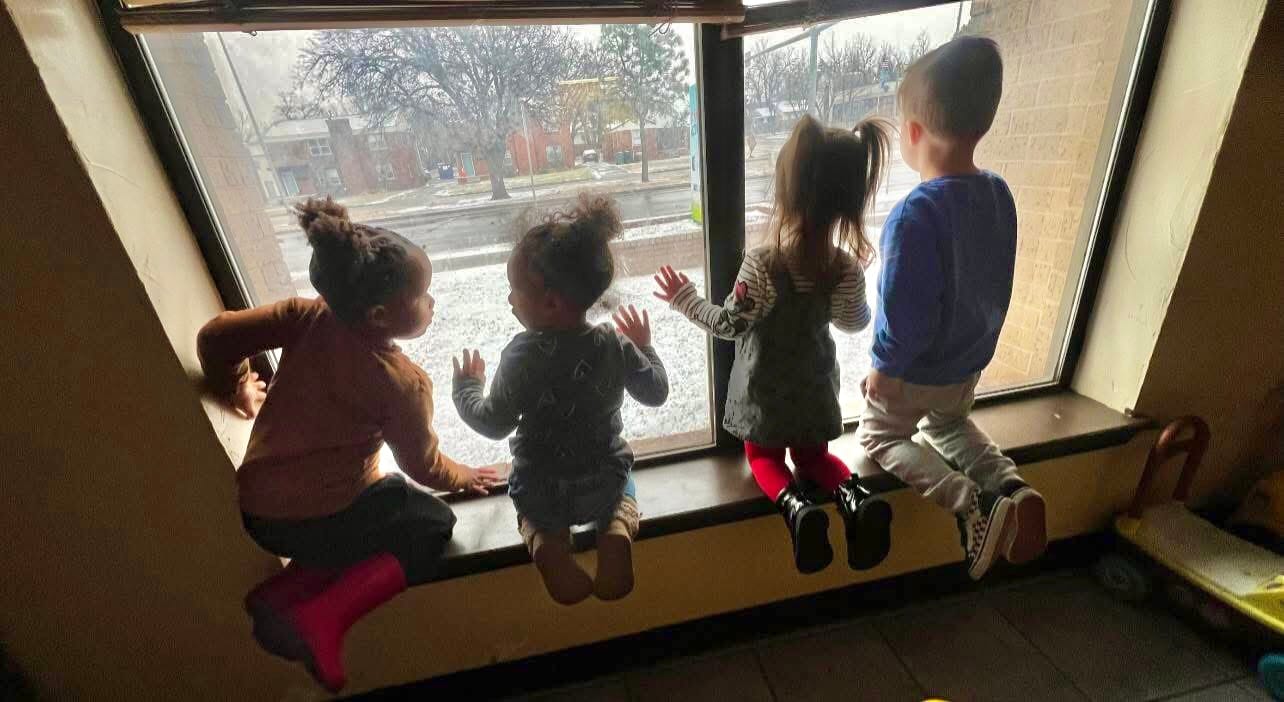 Four young children sitting on a windowsill, looking outside on a snowy day with curious expressions.